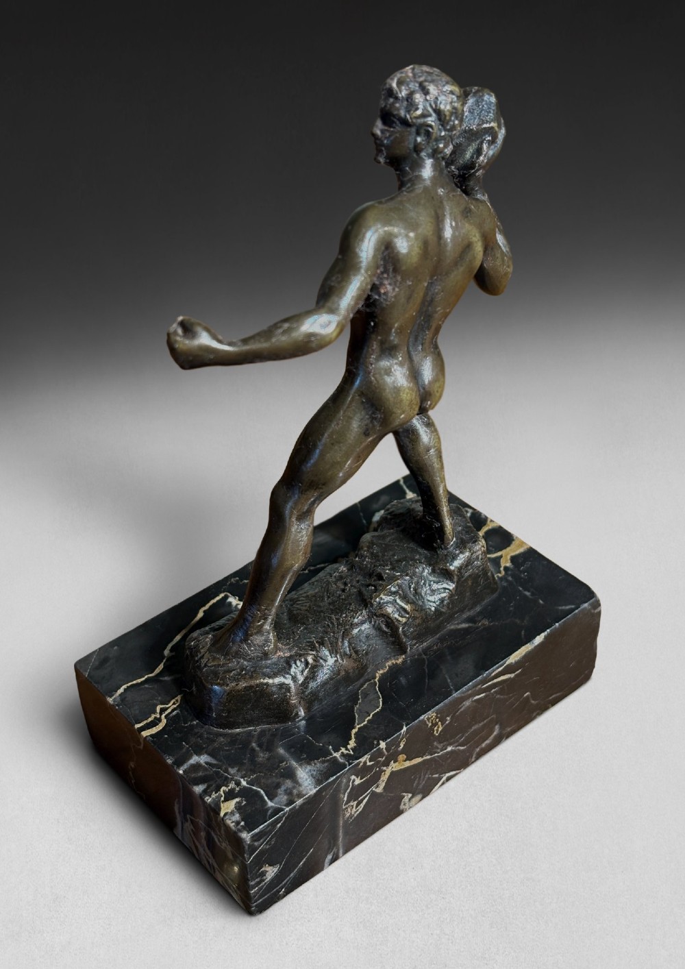 A patinated dark bronze figurine depicting a nude ancient Greek male athlete holding a stone, mounted on a rectangular black marble base with white veining. View II