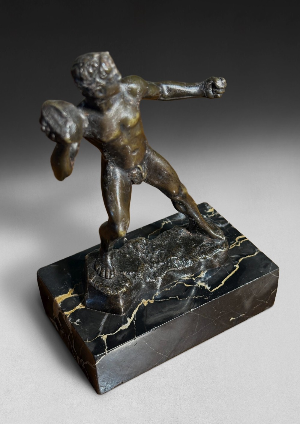 A patinated dark bronze figurine depicting a nude ancient Greek male athlete holding a stone, mounted on a rectangular black marble base with white veining. View IV