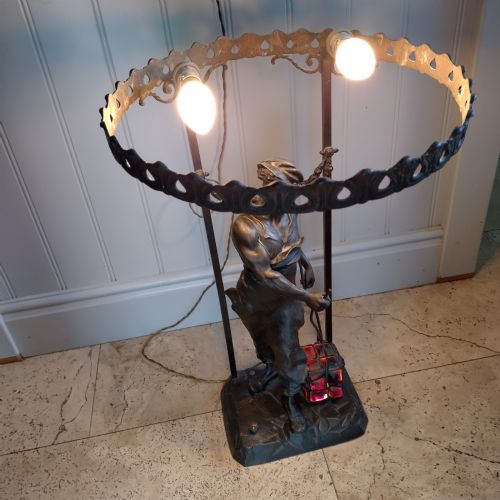 fig 1. A dark brown spelter table lamp featuring a sculpted figure of an industrial worker carrying a glowing red basket, topped with a perforated metal gallery ring holding light sockets.