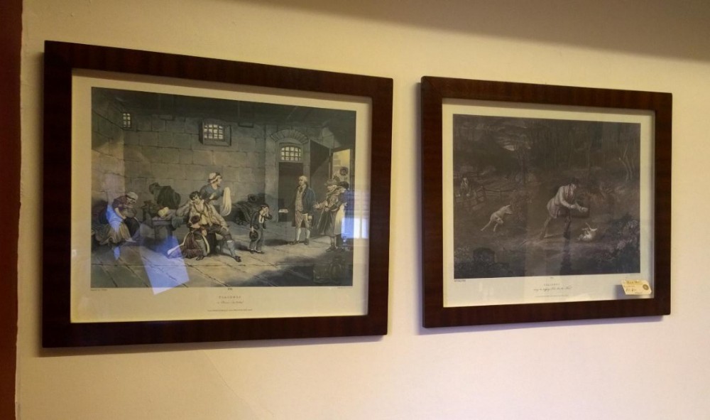 A pair of antique narrative engravings presented in matching rectangular dark mahogany frames hanging on a light wall.