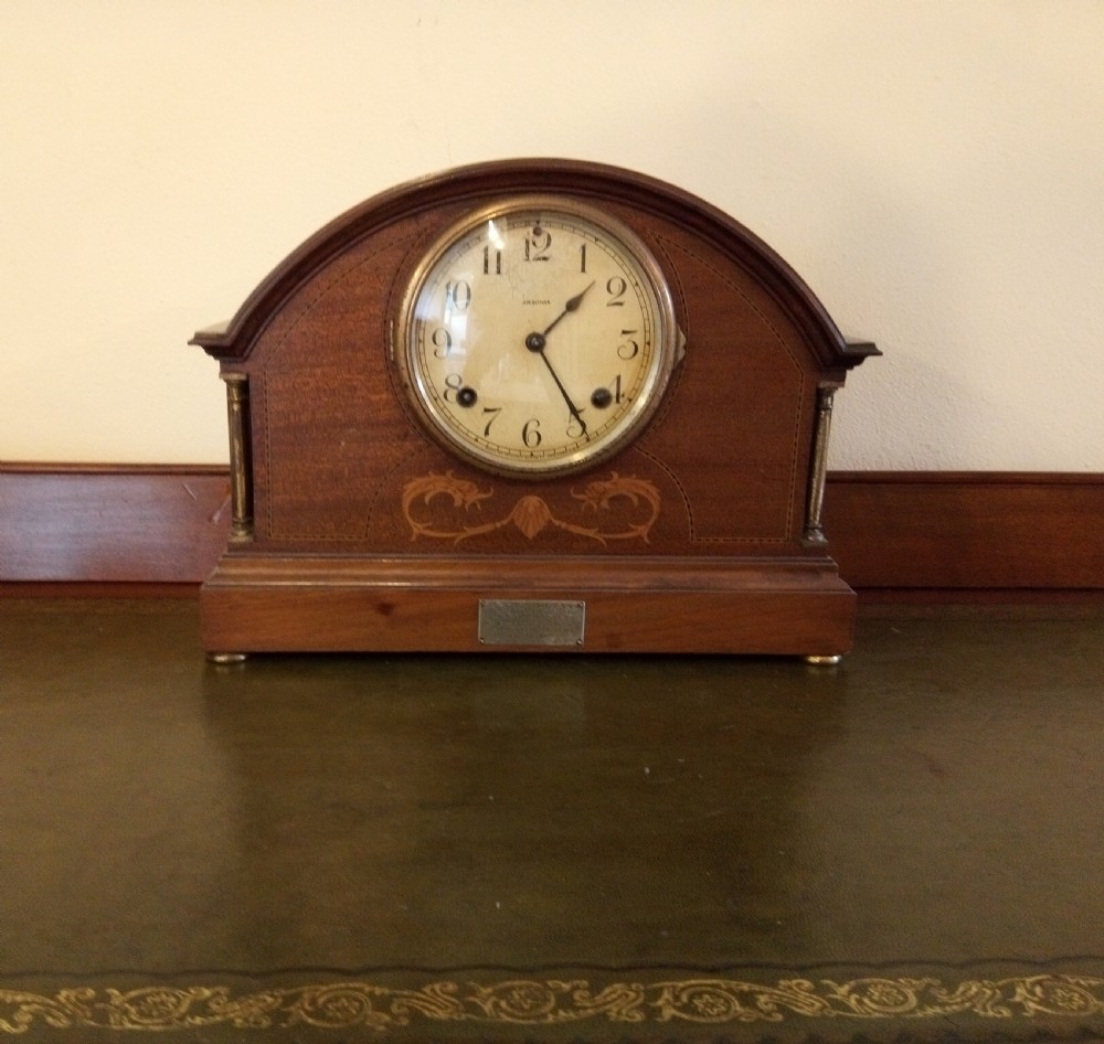 An Edwardian dome top mahogany mantel clock featuring floral marquetry inlay, brass side columns, and an Arabic numeral dial.