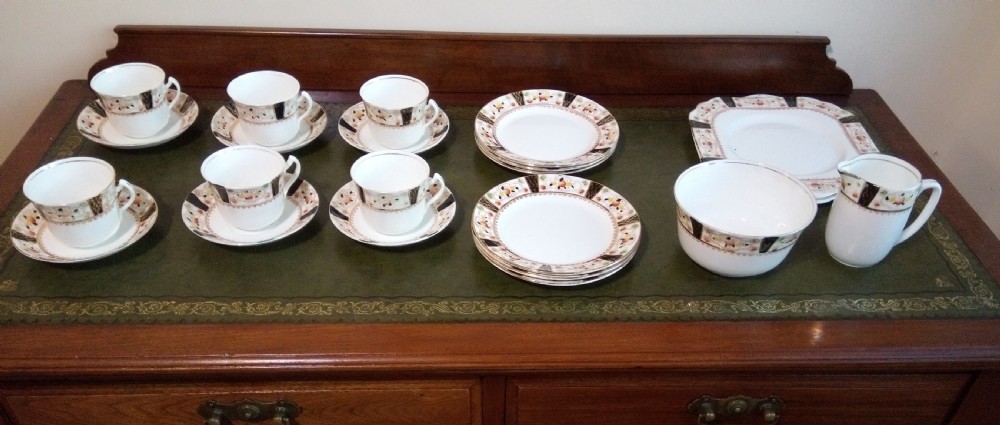 A twenty-one piece Gladstone bone china tea service arranged on a surface, displaying white teacups, saucers, side plates, a milk pitcher, a slop bowl, and a square serving plate decorated with black geometric borders and gilded rims.