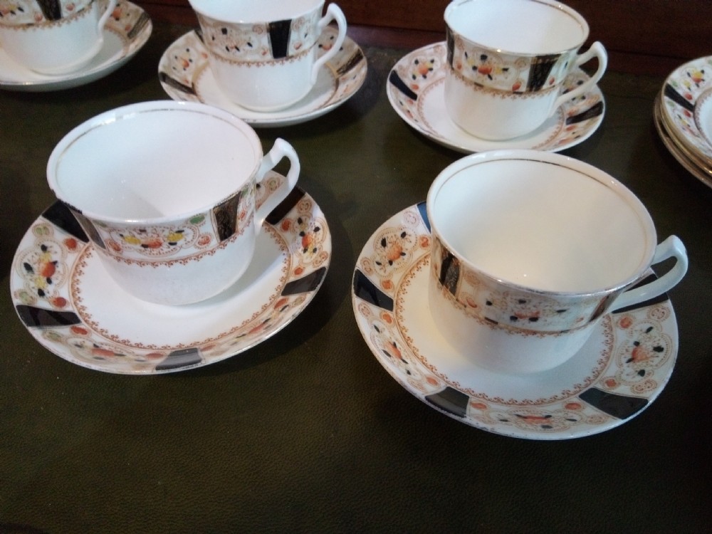 A twenty-one piece Gladstone bone china tea service arranged on a surface, displaying white teacups, saucers, side plates, a milk pitcher, a slop bowl, and a square serving plate decorated with black geometric borders and gilded rims. View II