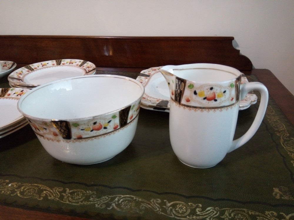 A twenty-one piece Gladstone bone china tea service arranged on a surface, displaying white teacups, saucers, side plates, a milk pitcher, a slop bowl, and a square serving plate decorated with black geometric borders and gilded rims. View III