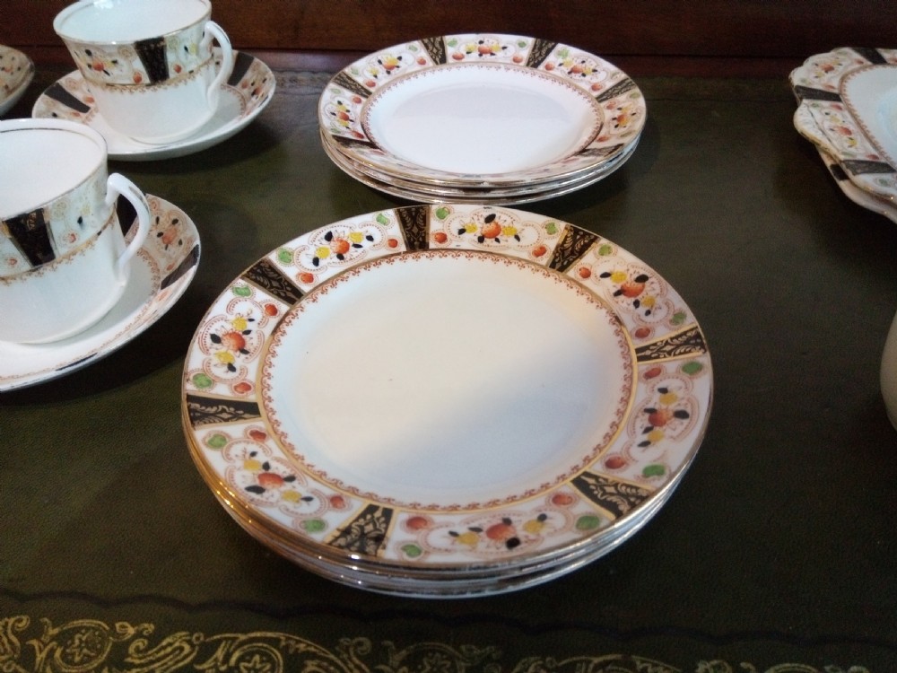 A twenty-one piece Gladstone bone china tea service arranged on a surface, displaying white teacups, saucers, side plates, a milk pitcher, a slop bowl, and a square serving plate decorated with black geometric borders and gilded rims. View VI