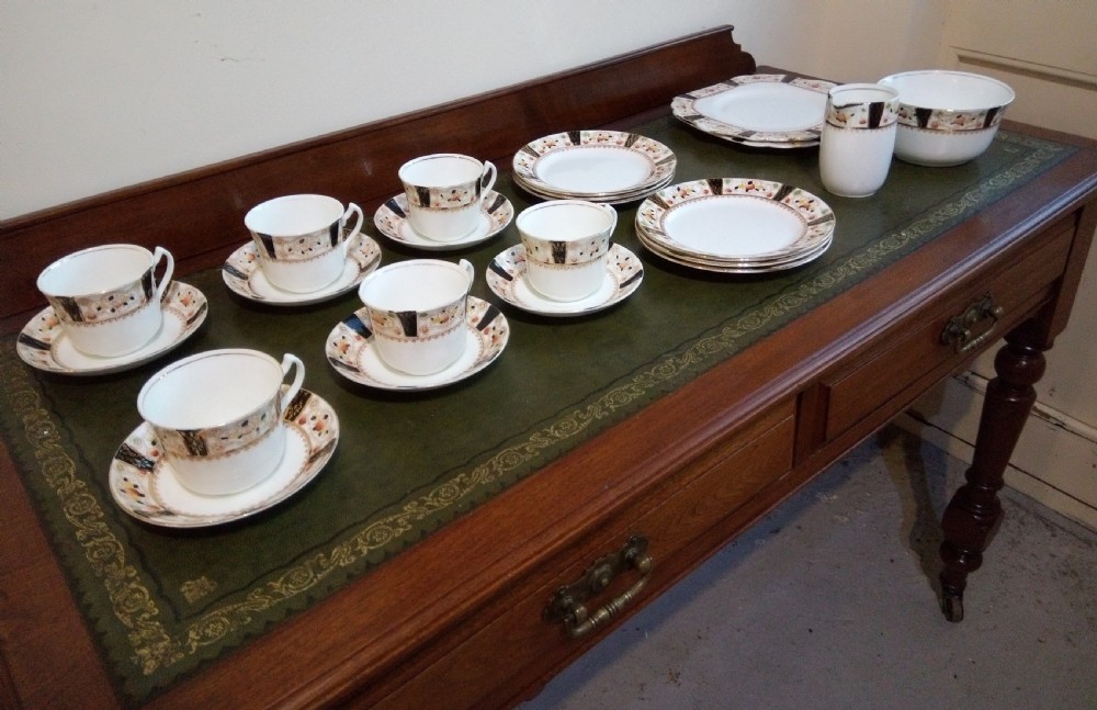 A twenty-one piece Gladstone bone china tea service arranged on a surface, displaying white teacups, saucers, side plates, a milk pitcher, a slop bowl, and a square serving plate decorated with black geometric borders and gilded rims. View VII