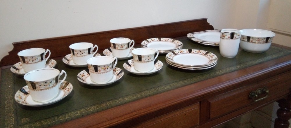 A twenty-one piece Gladstone bone china tea service arranged on a surface, displaying white teacups, saucers, side plates, a milk pitcher, a slop bowl, and a square serving plate decorated with black geometric borders and gilded rims. View VIII