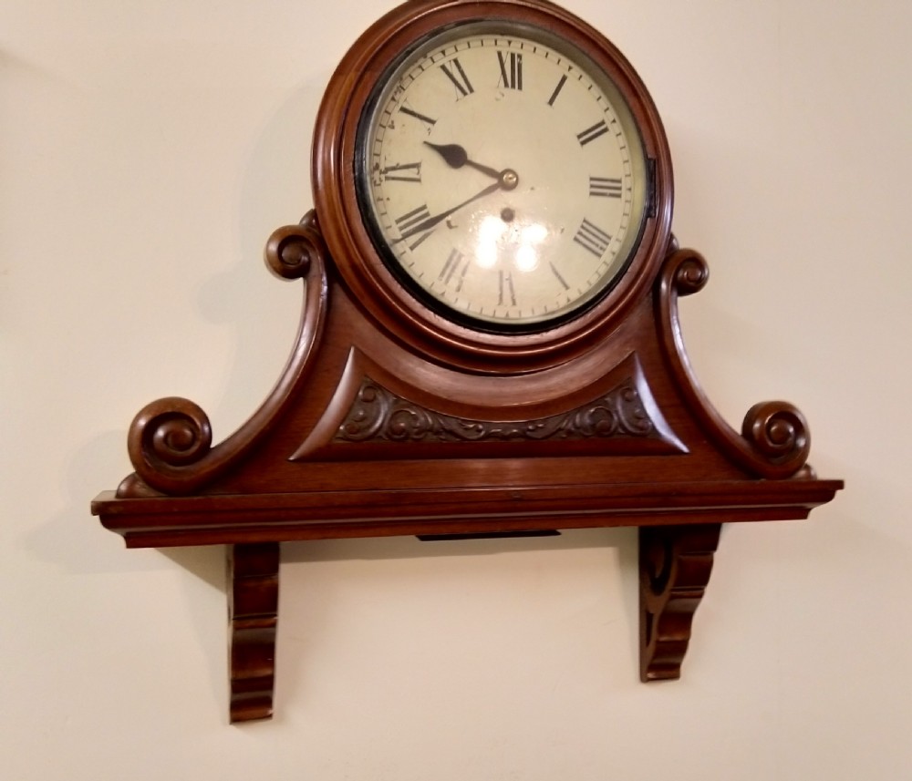 A 19th-century mahogany wall clock featuring a circular dial with Roman numerals resting on a carved wooden bracket support.