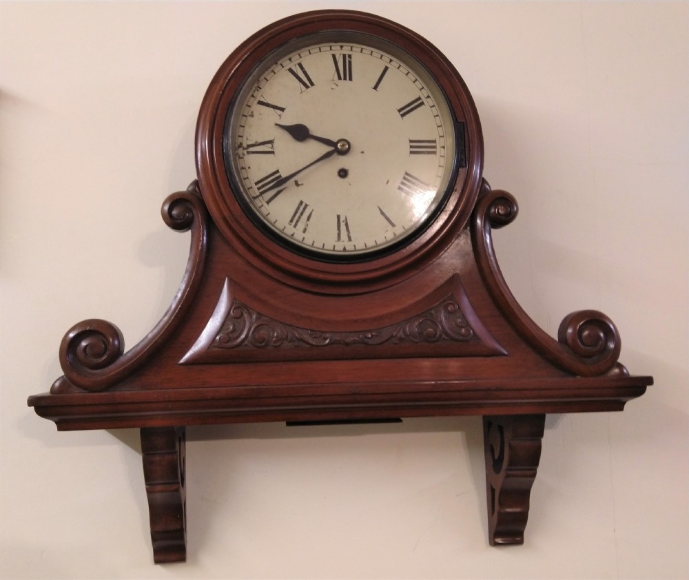 A 19th-century mahogany wall clock featuring a circular dial with Roman numerals resting on a carved wooden bracket support. View II