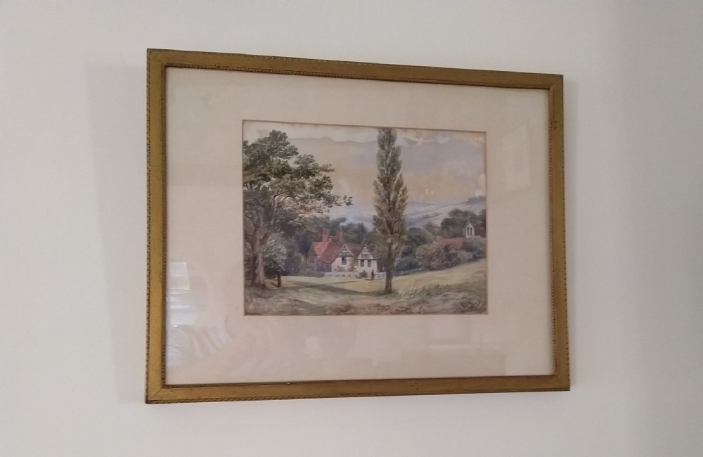 A framed watercolour painting depicting an English country landscape with a Tudor-style building and tall trees inside a cream mount and gold beaded frame.