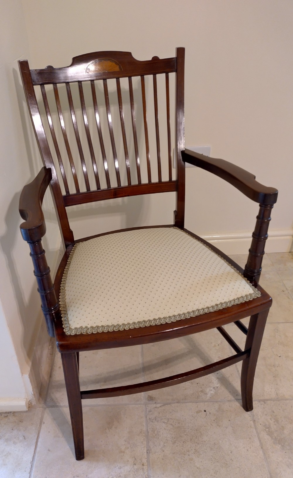 An Edwardian mahogany elbow chair featuring a vertical slat back with shell marquetry inlay, shaped arms, and a cream upholstered seat.