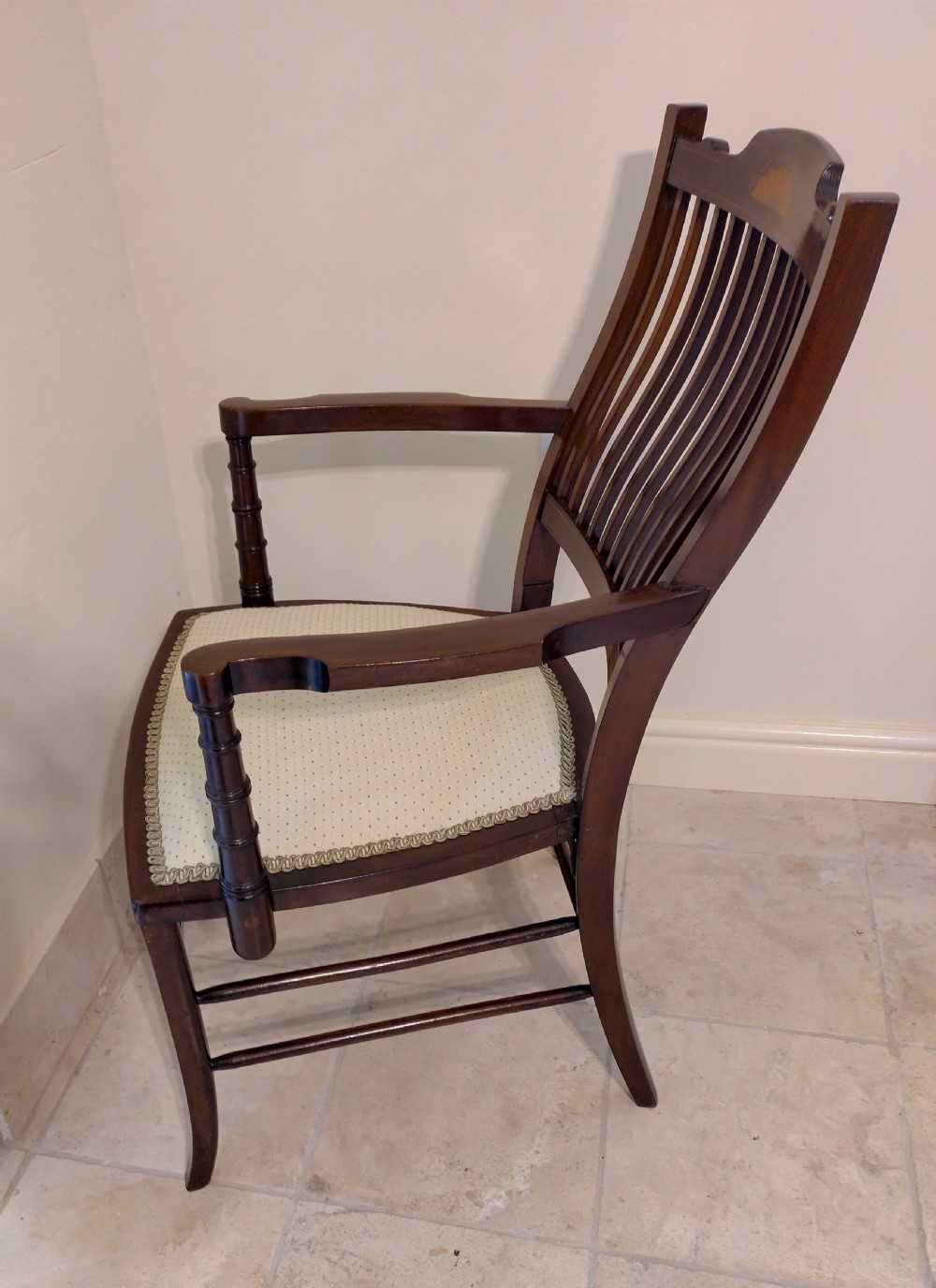 An Edwardian mahogany elbow chair featuring a vertical slat back with shell marquetry inlay, shaped arms, and a cream upholstered seat. View VI