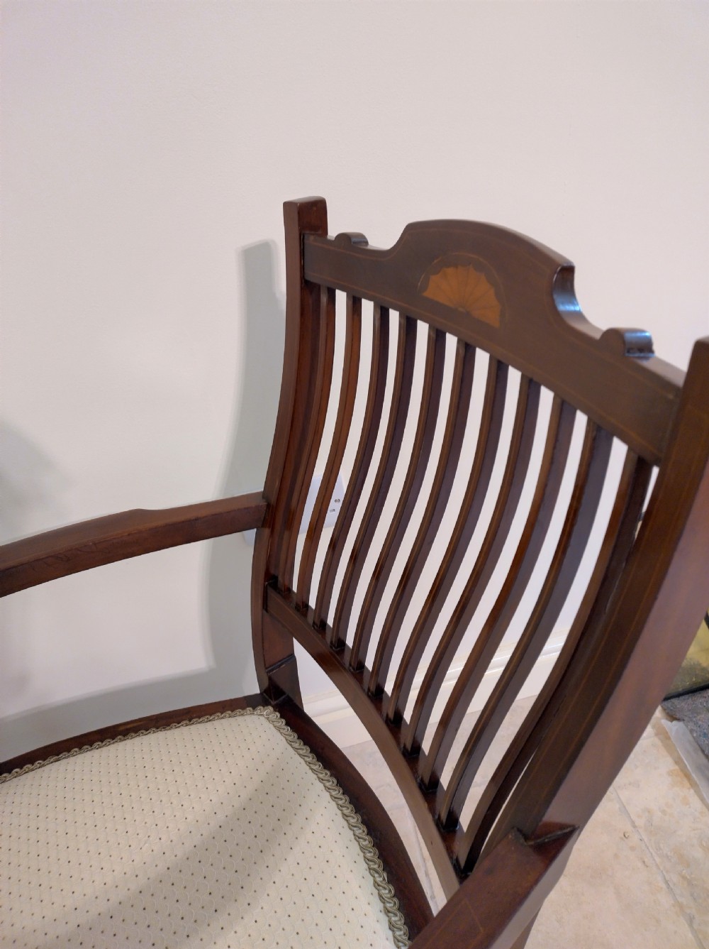 An Edwardian mahogany elbow chair featuring a vertical slat back with shell marquetry inlay, shaped arms, and a cream upholstered seat. View VII