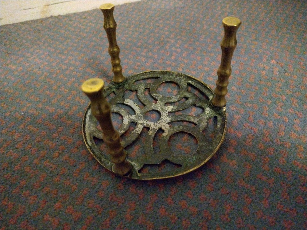 A circular cast brass trivet with a pierced geometric pattern top supported by four turned legs. View IV