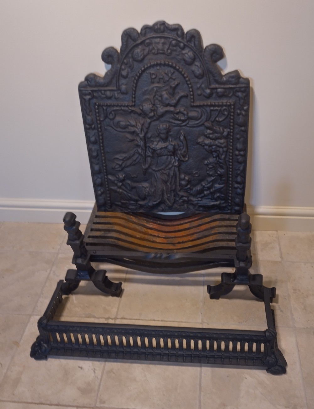A heavy black cast iron fireback featuring pictorial relief carving, mounted above a matching fire grate with an accompanying low pierced iron fender curb.