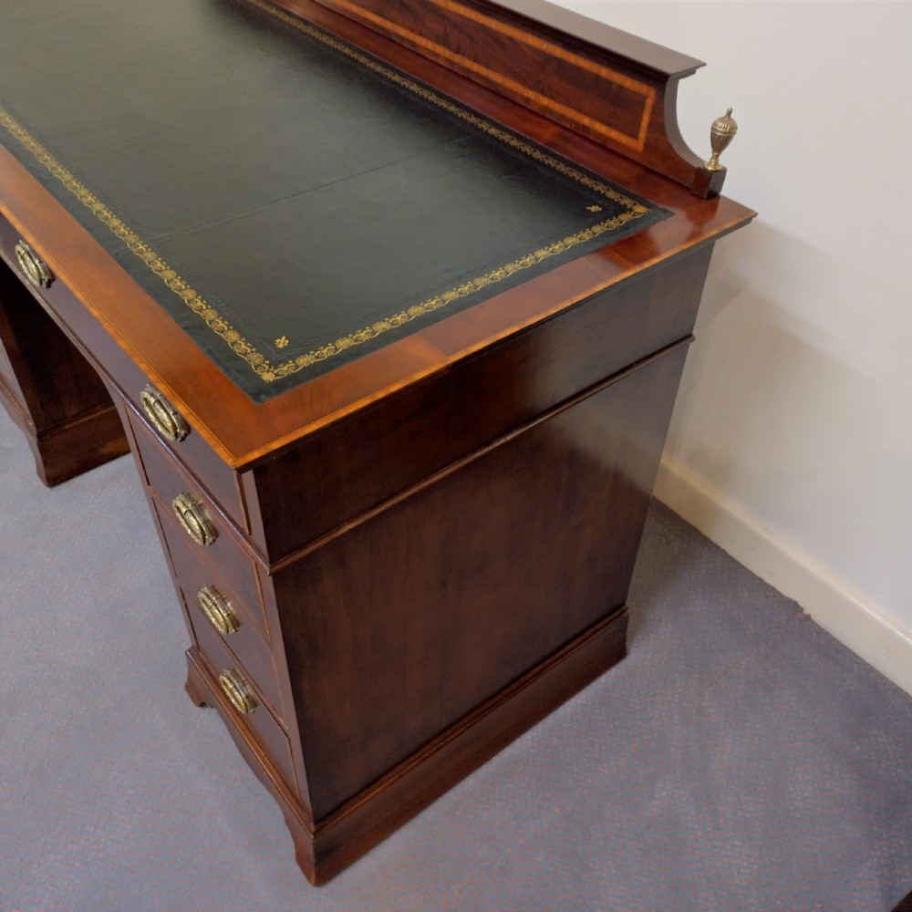 An Edwardian inlaid mahogany pedestal desk featuring twin pedestals with multiple drawers, brass oval drop handles, a dark leather writing insert, and a low gallery back with urn finials. View VII