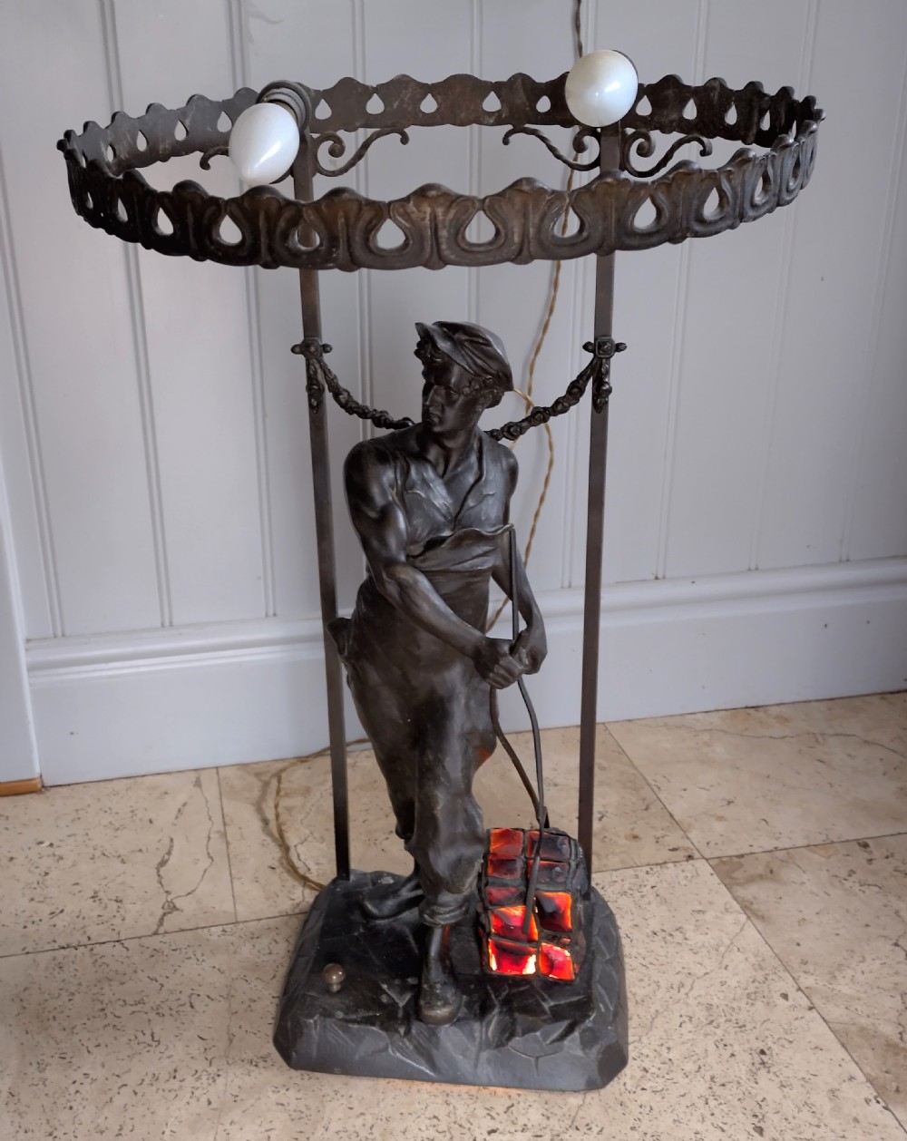 A dark brown spelter table lamp featuring a sculpted figure of an industrial worker carrying a glowing red basket, topped with a perforated metal gallery ring holding light sockets.