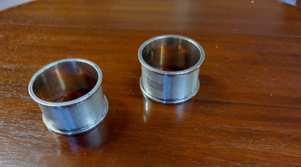 A pair of cylindrical silver napkin rings with engine-turned geometric textures and plain rectangular cartouches resting on a polished wooden surface.