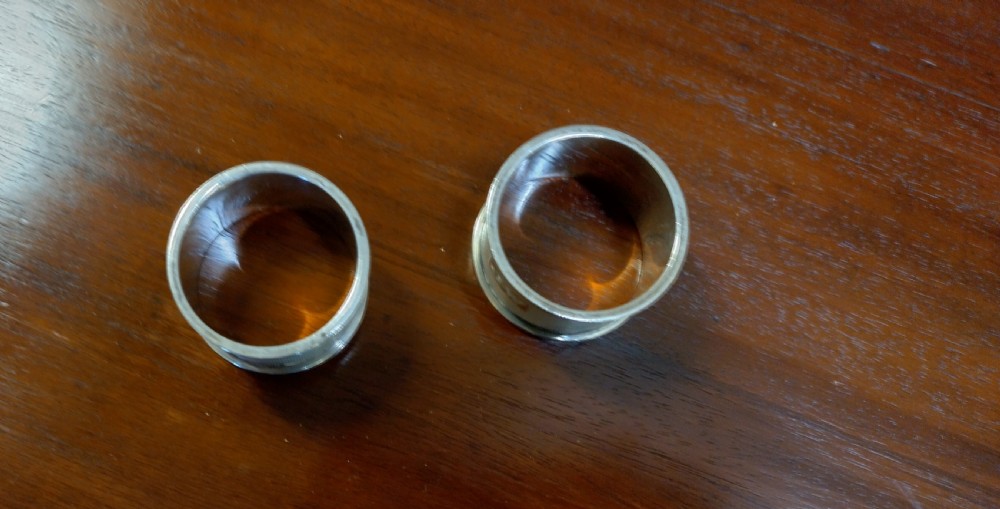 A pair of cylindrical silver napkin rings with engine-turned geometric textures and plain rectangular cartouches resting on a polished wooden surface. View II