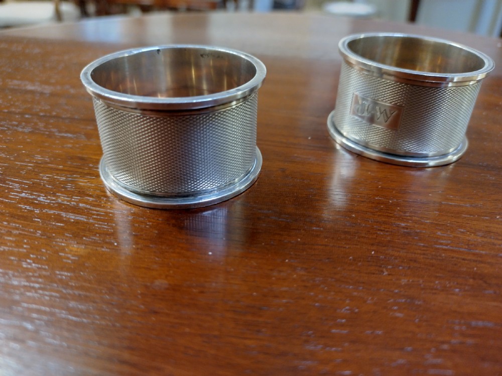 A pair of cylindrical silver napkin rings with engine-turned geometric textures and plain rectangular cartouches resting on a polished wooden surface. View III