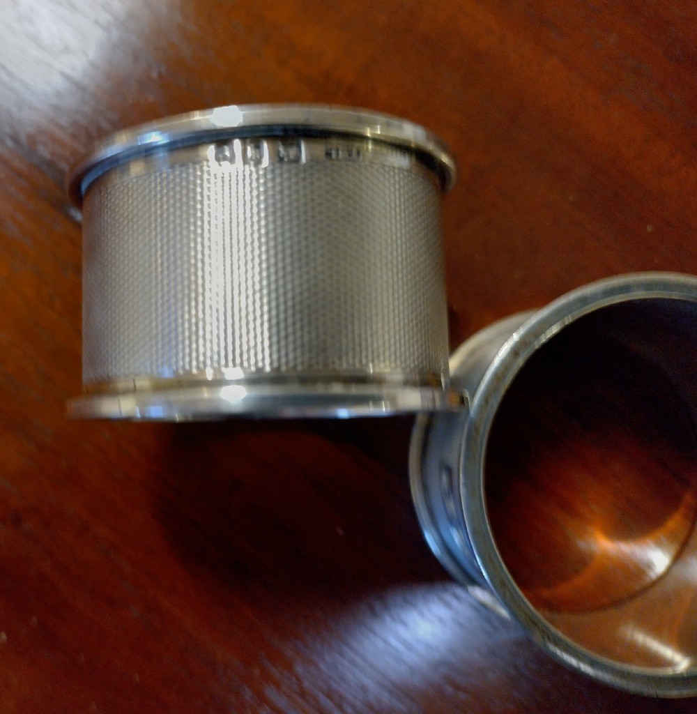 A pair of cylindrical silver napkin rings with engine-turned geometric textures and plain rectangular cartouches resting on a polished wooden surface. View IV