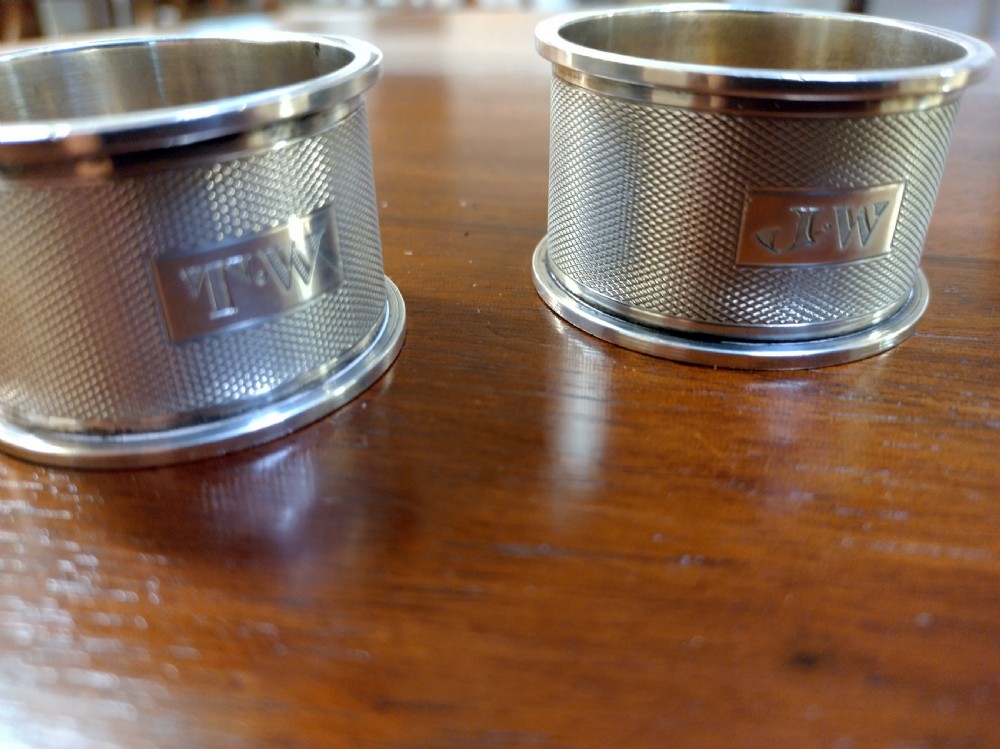 A pair of cylindrical silver napkin rings with engine-turned geometric textures and plain rectangular cartouches resting on a polished wooden surface. View V