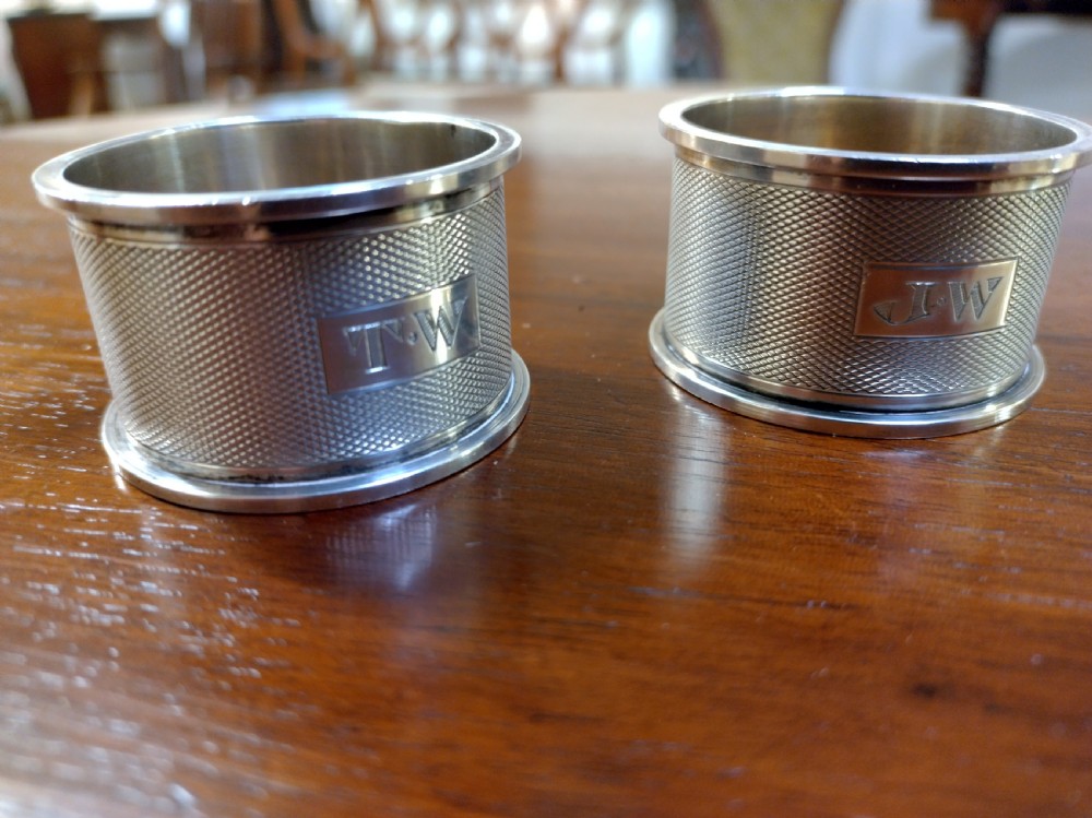 A pair of cylindrical silver napkin rings with engine-turned geometric textures and plain rectangular cartouches resting on a polished wooden surface. View VI