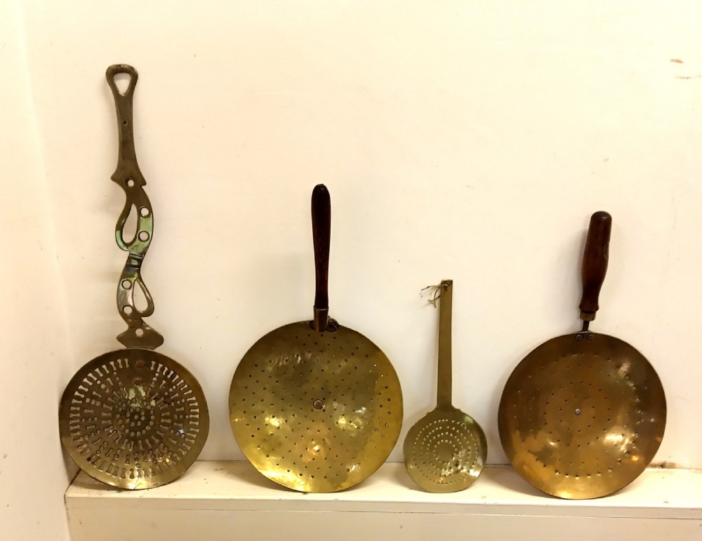 A collection of four antique brass cream skimmers with pierced round bowls, featuring turned wooden handles and decorative flat brass handles.