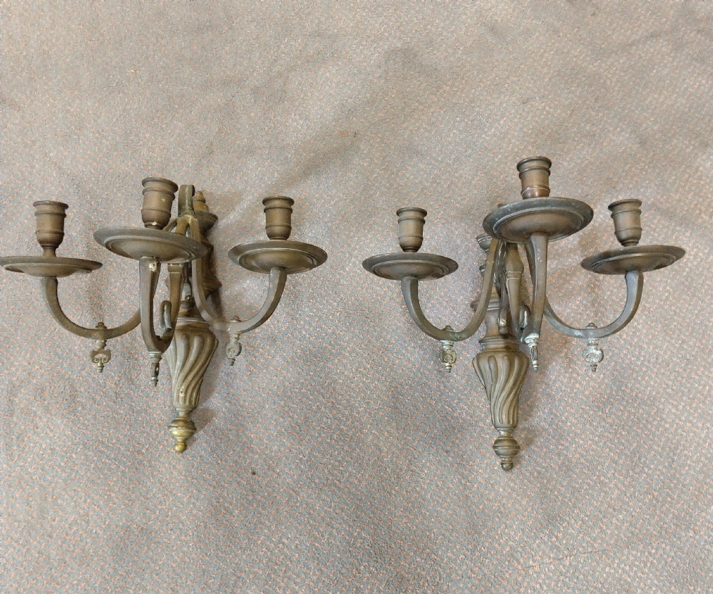 A pair of patinated three-arm solid brass wall sconces featuring fluted urn-shaped backplates and curved candle branches.