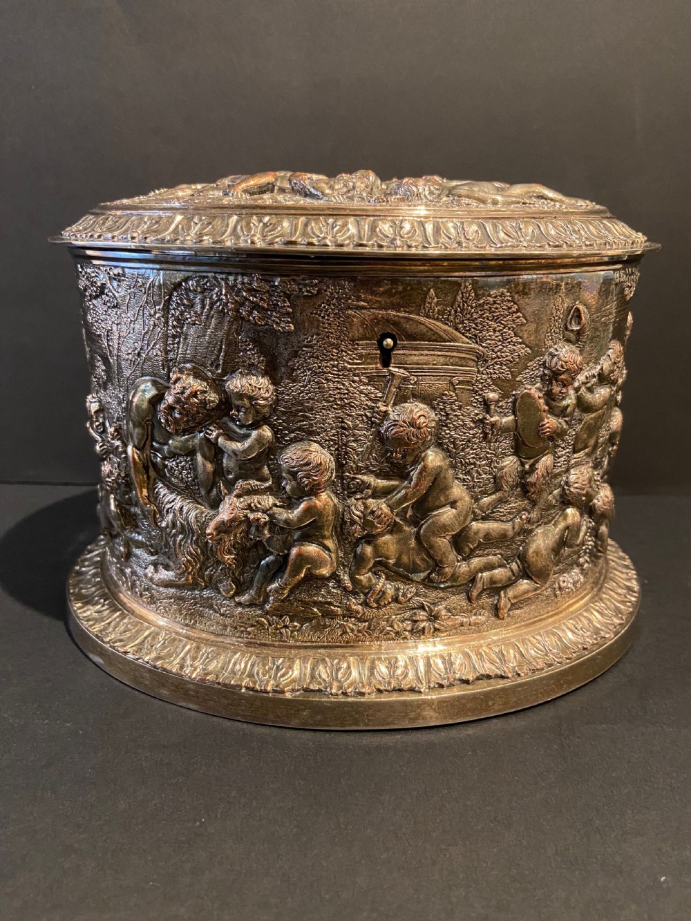 An oval silver-plated tea caddy decorated with high-relief raised figures of cherubs in a landscape setting with gadrooned trim around the base and lid.