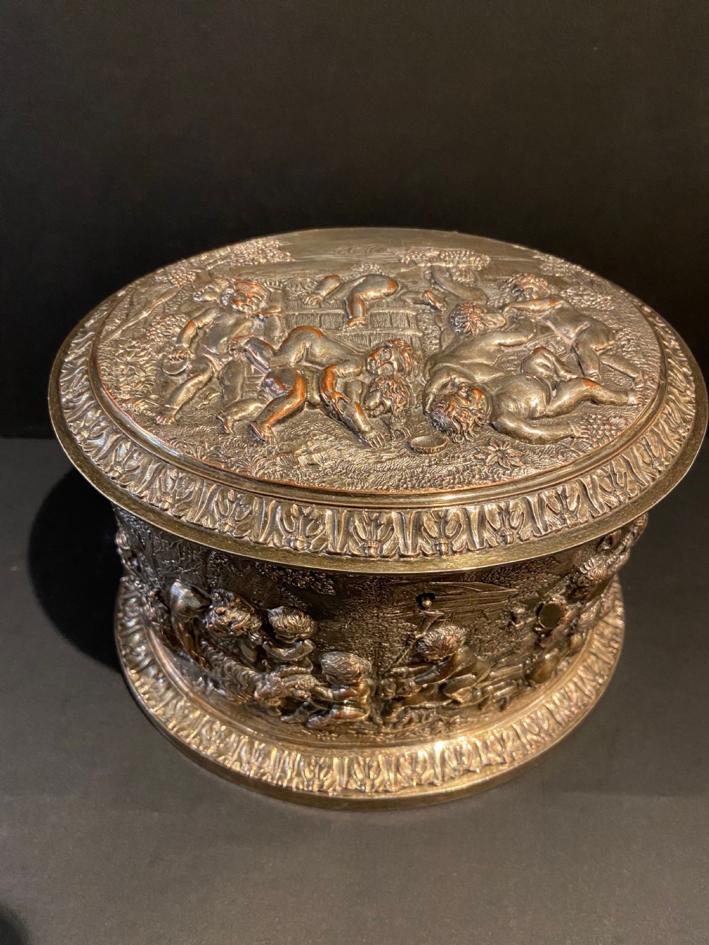 An oval silver-plated tea caddy decorated with high-relief raised figures of cherubs in a landscape setting with gadrooned trim around the base and lid. View II