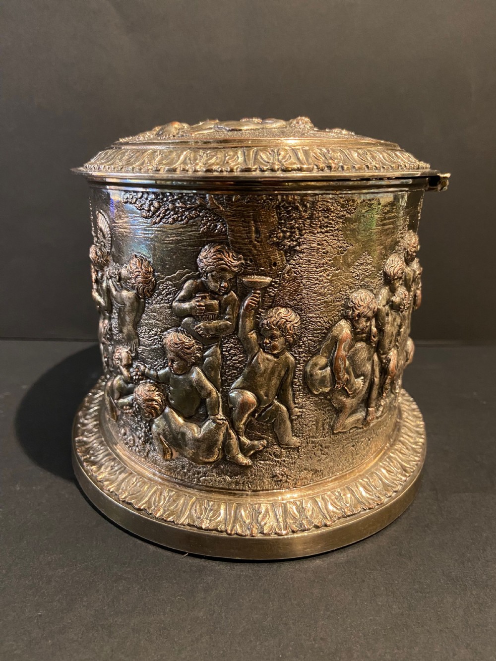 An oval silver-plated tea caddy decorated with high-relief raised figures of cherubs in a landscape setting with gadrooned trim around the base and lid. View III