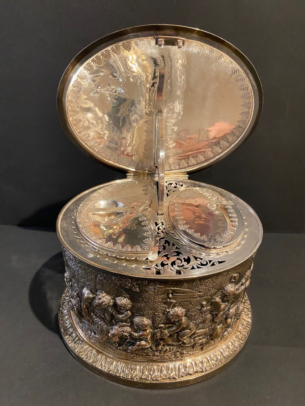 An oval silver-plated tea caddy decorated with high-relief raised figures of cherubs in a landscape setting with gadrooned trim around the base and lid. View IV