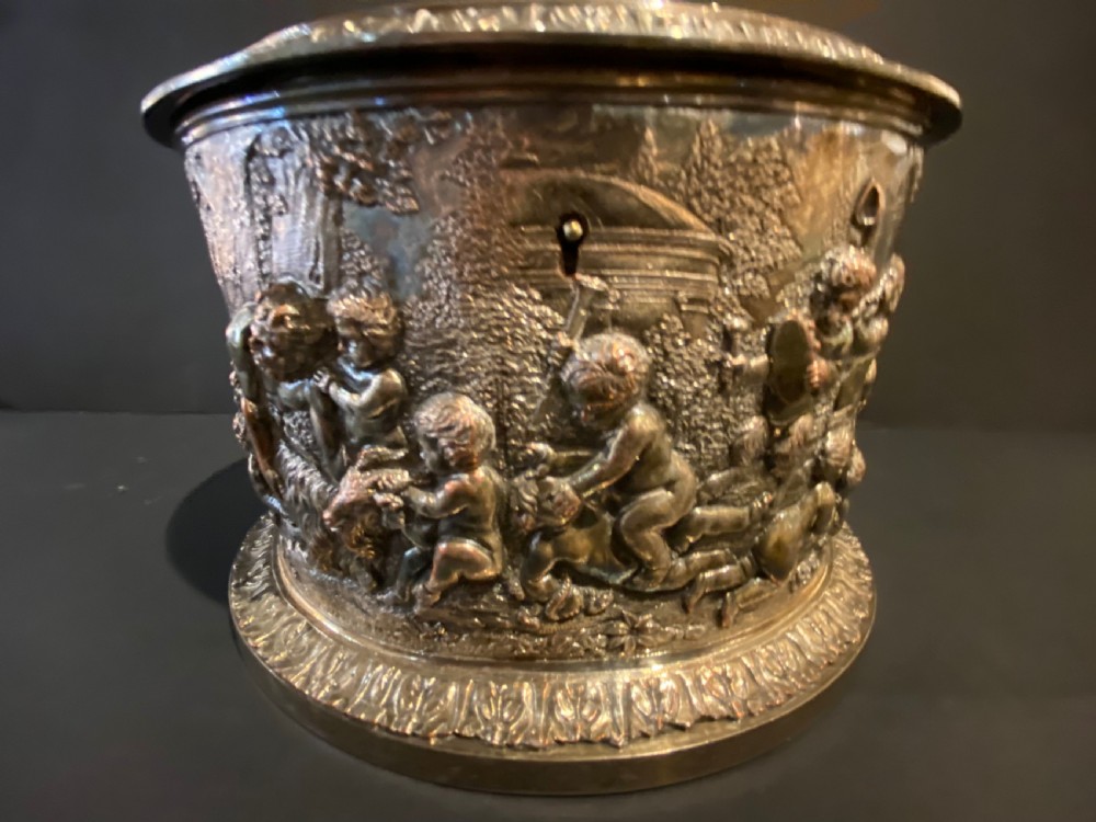 An oval silver-plated tea caddy decorated with high-relief raised figures of cherubs in a landscape setting with gadrooned trim around the base and lid. View VI