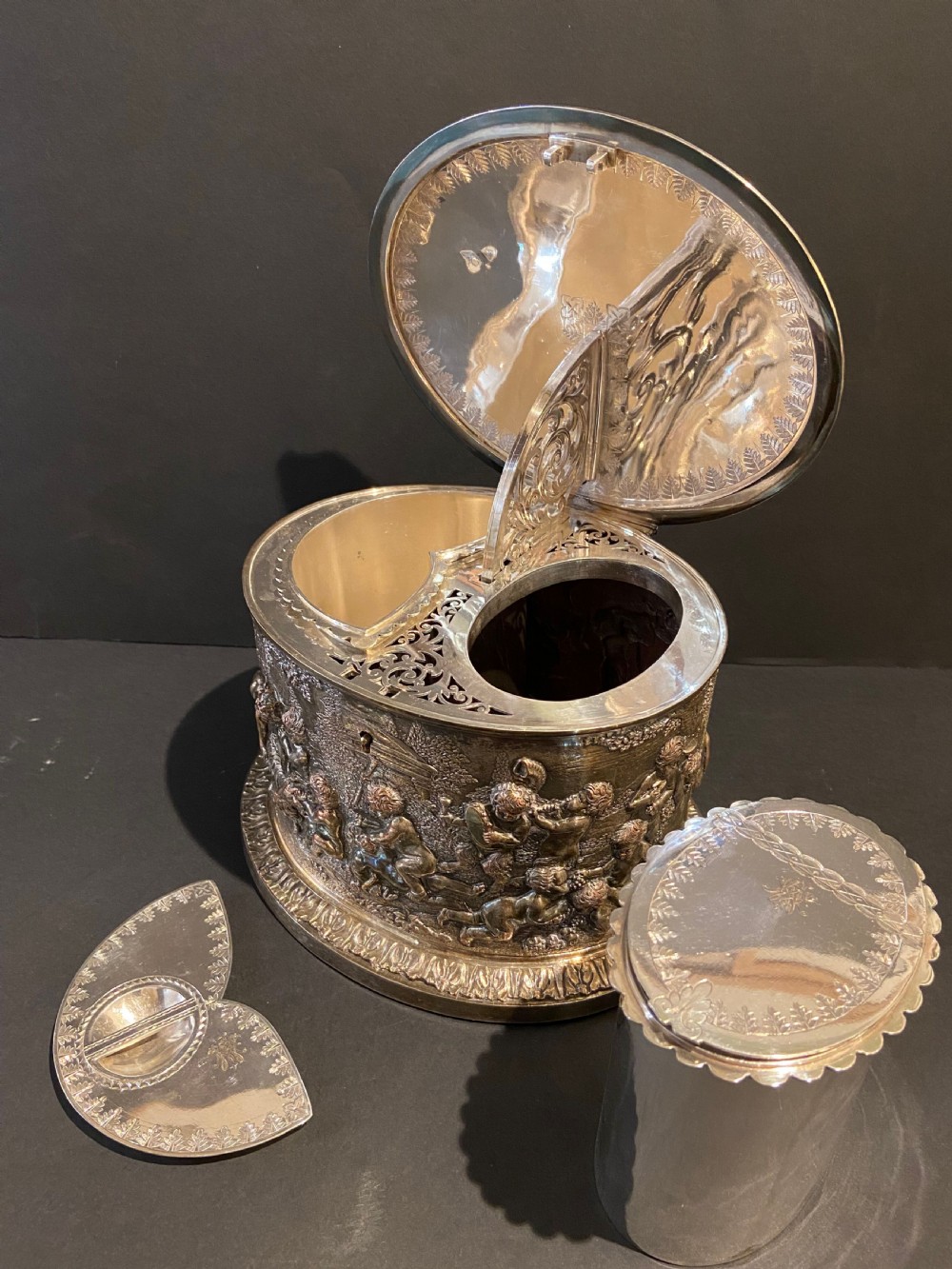 An oval silver-plated tea caddy decorated with high-relief raised figures of cherubs in a landscape setting with gadrooned trim around the base and lid. View VII