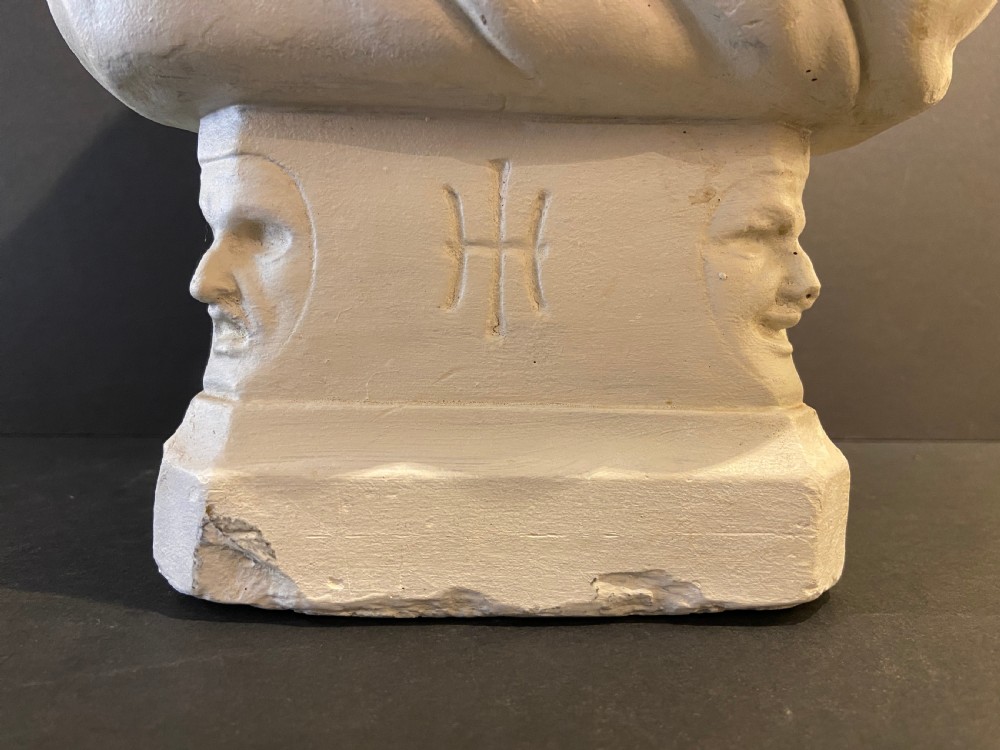 An off-white plaster portrait bust depicting a male figure with draped classical fabric, set upon a square plinth base with carved motifs. View II