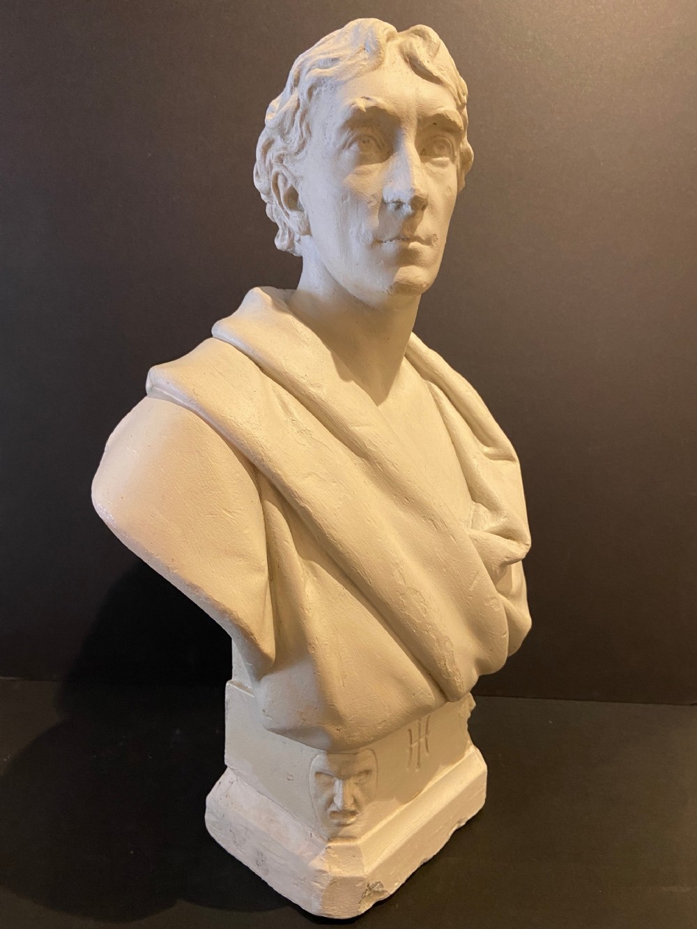 An off-white plaster portrait bust depicting a male figure with draped classical fabric, set upon a square plinth base with carved motifs.