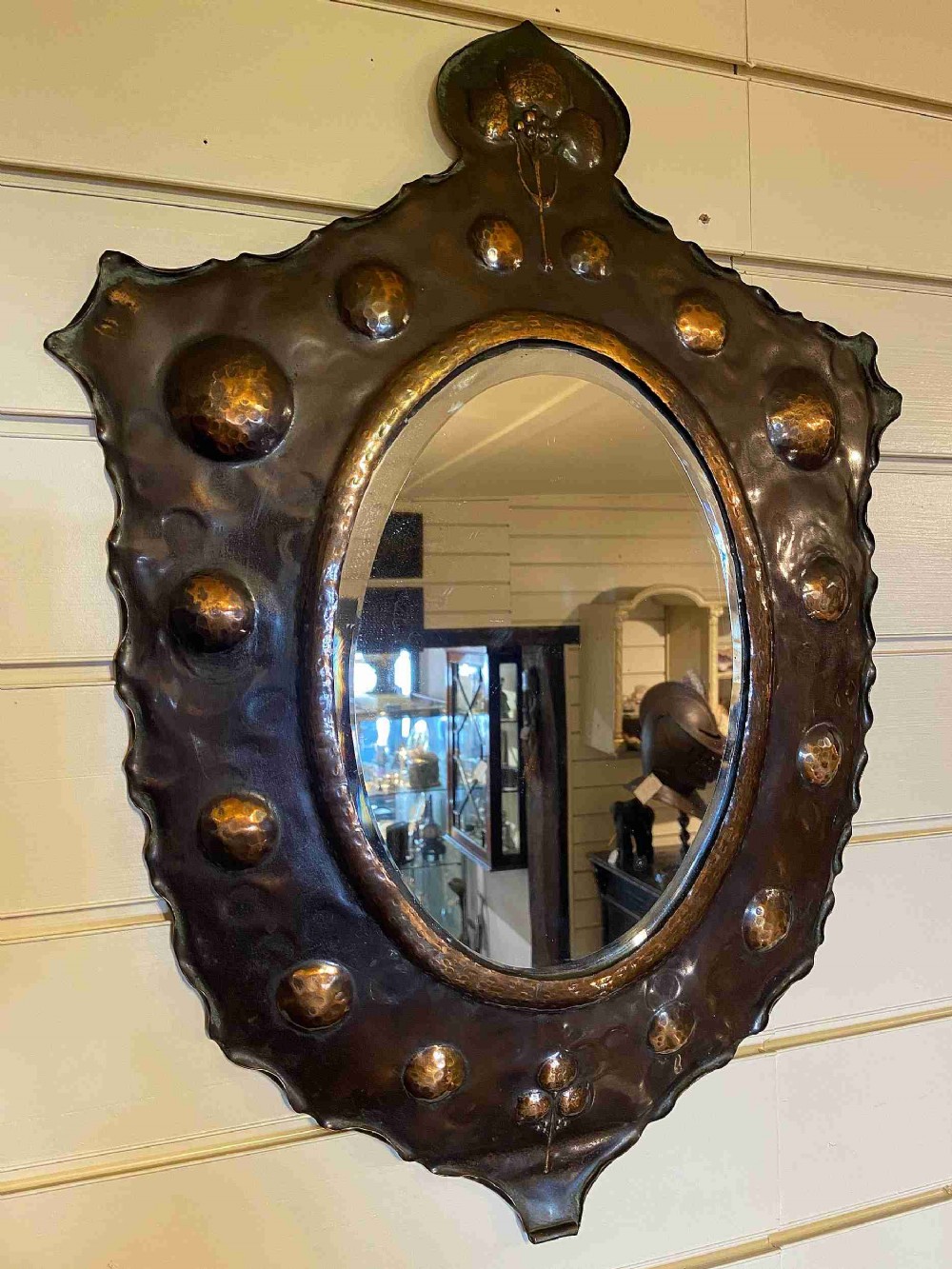 A late 19th-century Arts and Crafts oval wall mirror featuring a hand-hammered patinated copper frame decorated with raised bosses and stylised floral motifs.