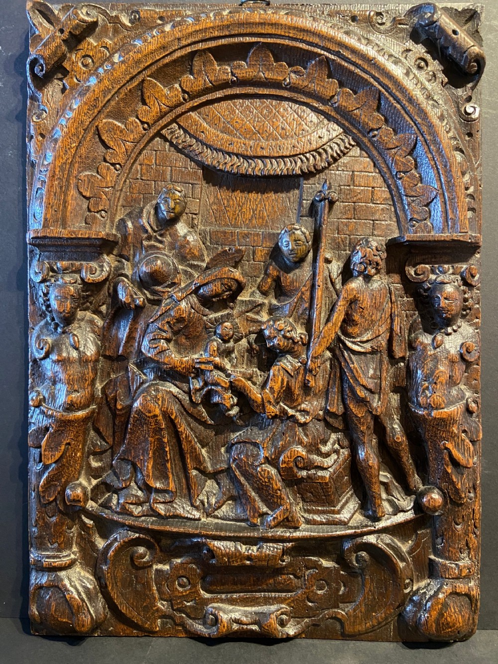 A 16th-century dark oak carved wood panel featuring a high-relief biblical scene enclosed within an arched frame supported by carved side columns.