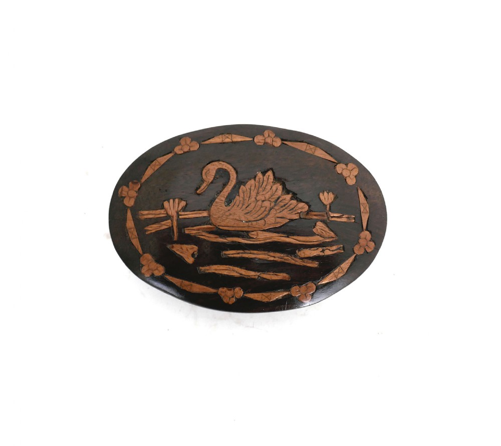 An oval dark lignum vitae wooden paperweight featuring lighter boxwood inlay depicting a swan floating on water surrounded by a decorative border.