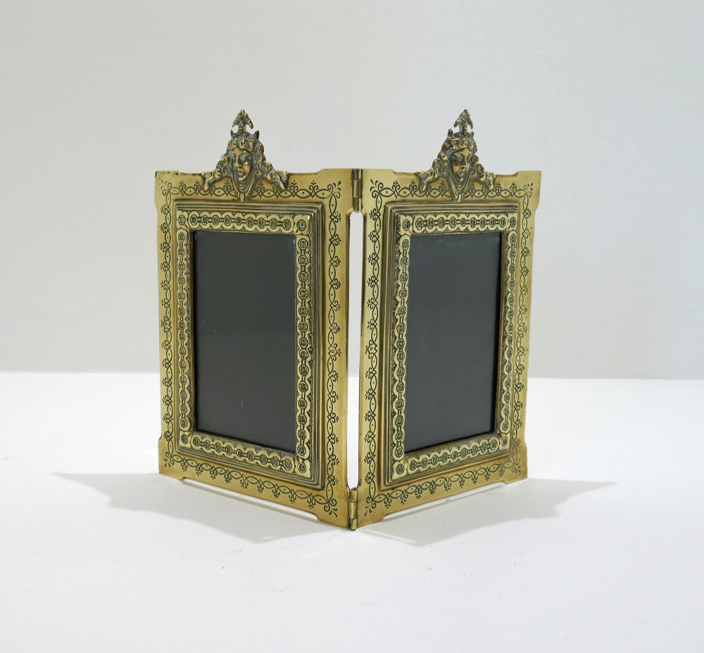 A late 19th-century Victorian double brass picture frame with ornate engraved geometric borders and classical mask finials.