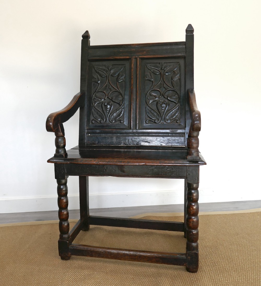 A 17th-century dark oak wainscot chair with carved floral back panels, turned front legs, and wooden perimeter stretchers.