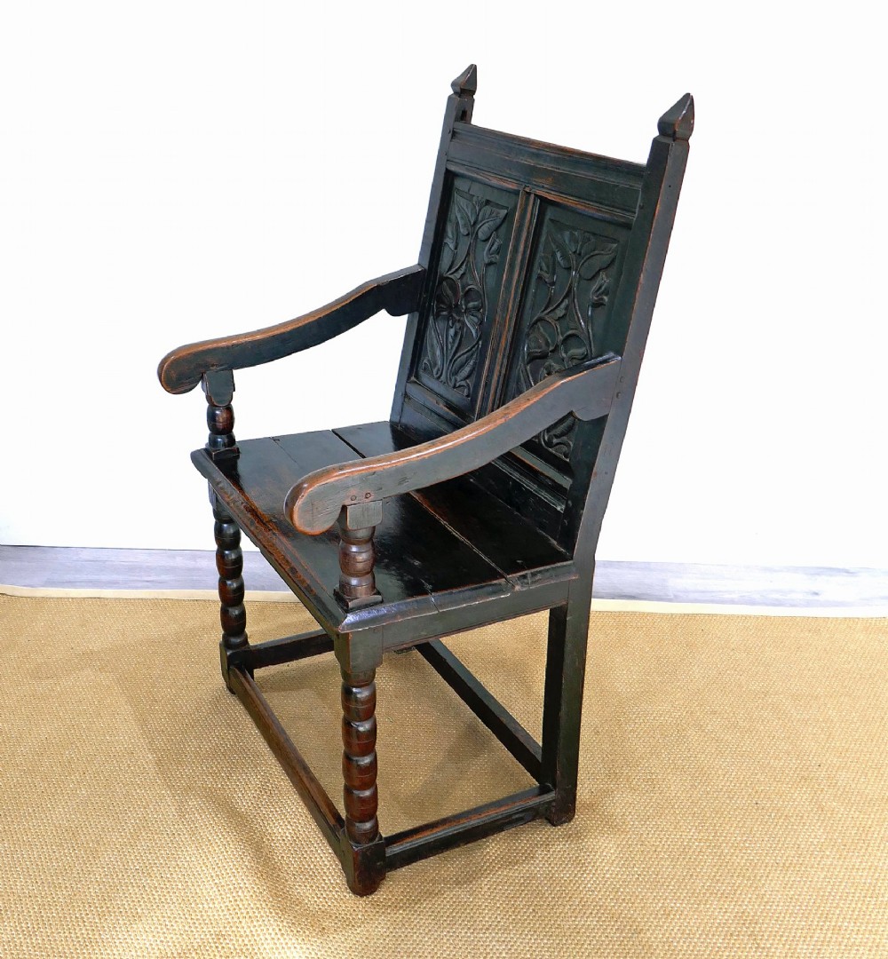 A 17th-century dark oak wainscot chair with carved floral back panels, turned front legs, and wooden perimeter stretchers. View V