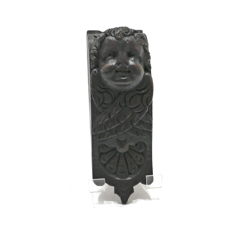 A dark carved oak architectural applique featuring a sculpted cherub head with folded wings and a fan motif at the base.