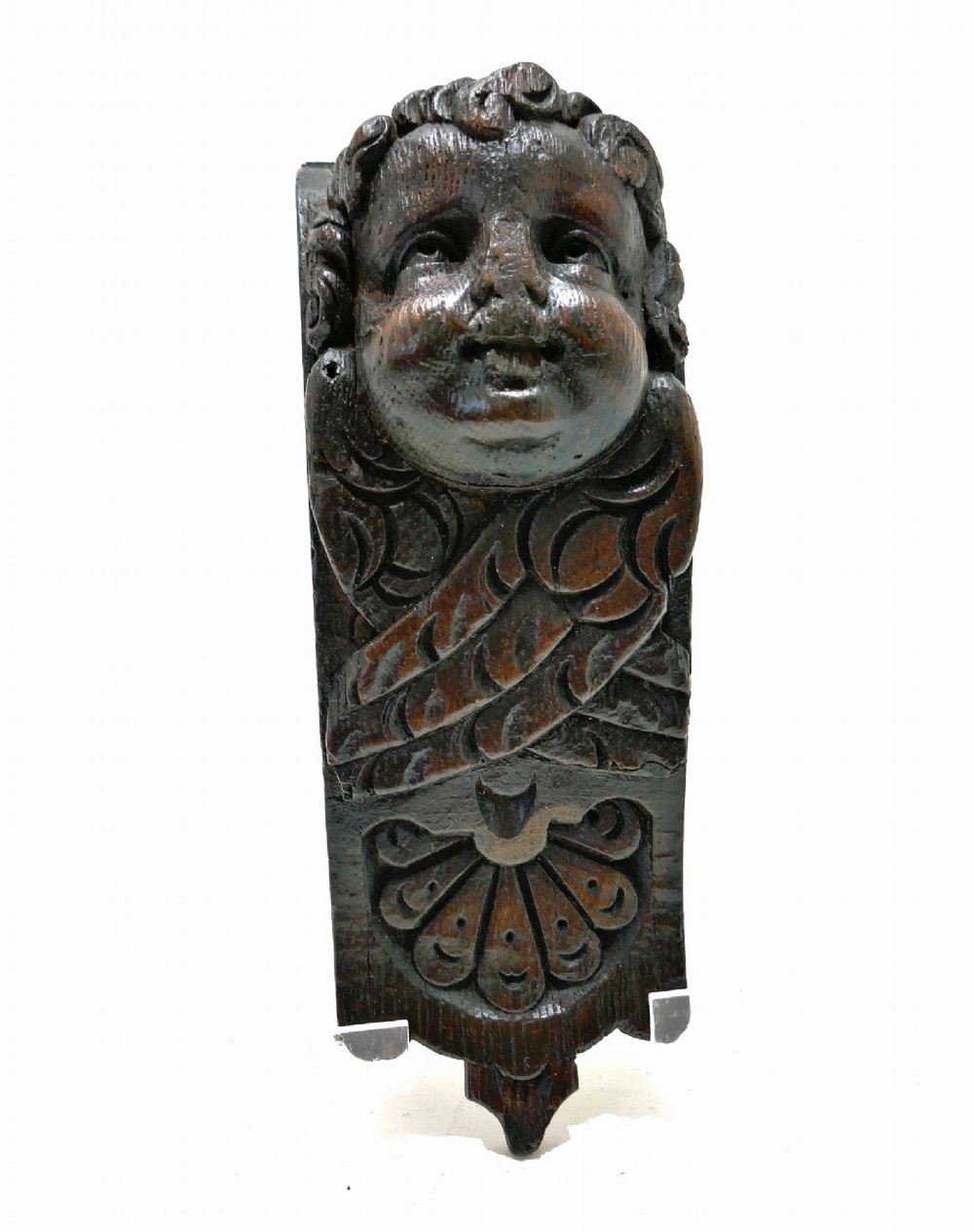 A dark carved oak architectural applique featuring a sculpted cherub head with folded wings and a fan motif at the base. View V