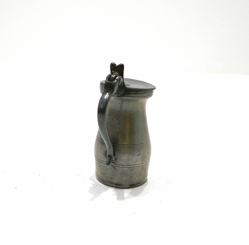 An 18th-century English pewter spirit measure featuring a flat hinged lid, curved handle, and incised horizontal lines around a baluster body. View VI