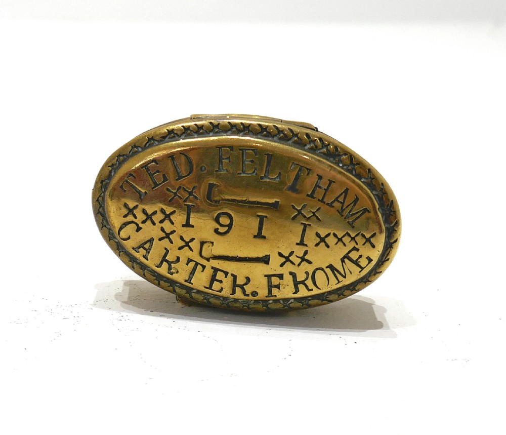An oval brass snuff box featuring hand-engraved text that reads Ted Feltham 1911 Carter Frome surrounded by small hammer motifs and decorative border crosshatching.