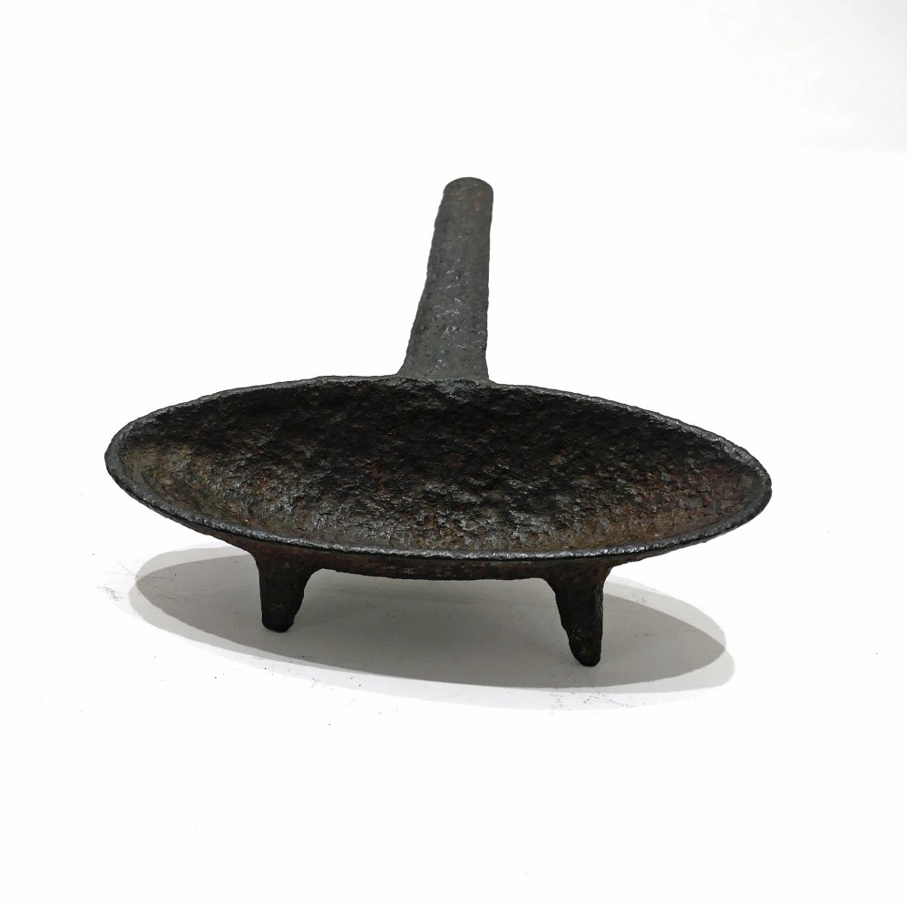 A 17th-century cast iron hearth grisset featuring a shallow boat-shaped vessel with three short tripod feet and a straight handle against a white backdrop.