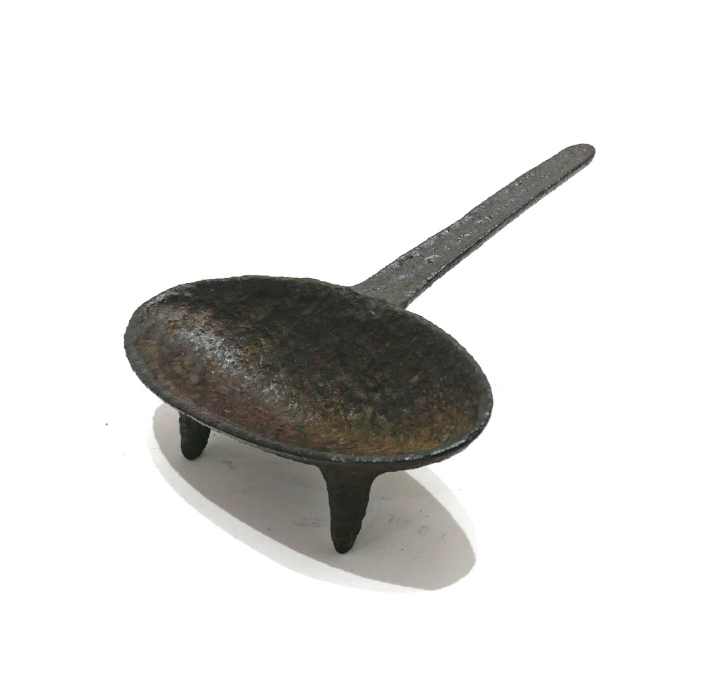 A 17th-century cast iron hearth grisset featuring a shallow boat-shaped vessel with three short tripod feet and a straight handle against a white backdrop. View II