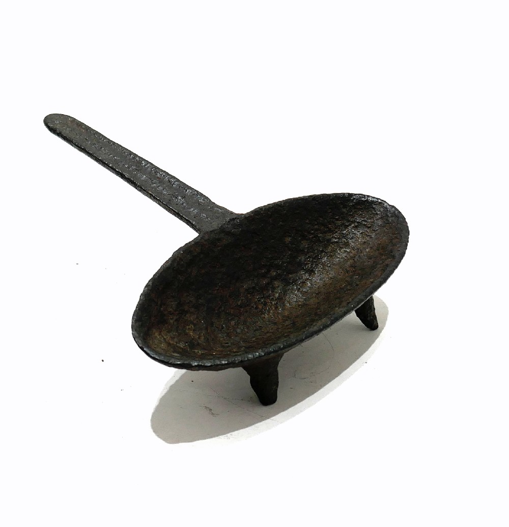 A 17th-century cast iron hearth grisset featuring a shallow boat-shaped vessel with three short tripod feet and a straight handle against a white backdrop. View III